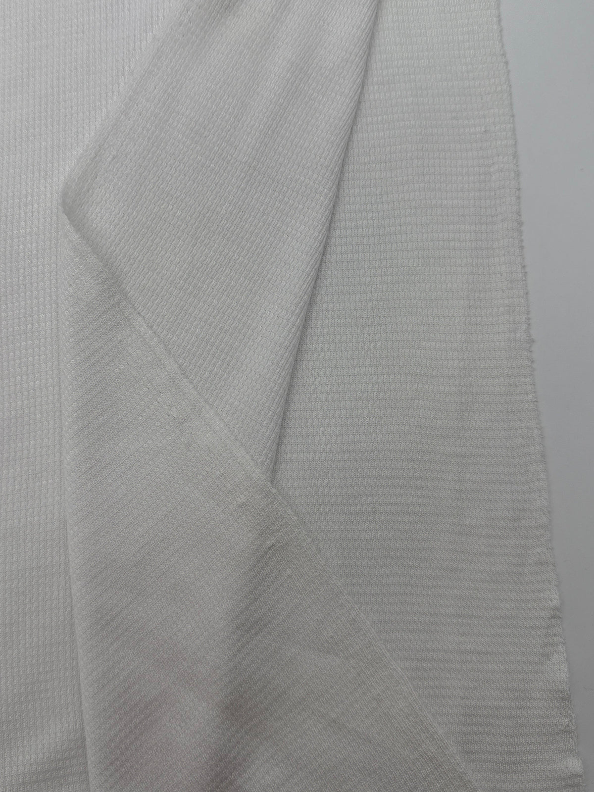 A close-up of a white ribbed knit fabric, likely a lightweight sweater or cardigan, showing the fine textured weave and folded seams against a neutral light gray background.