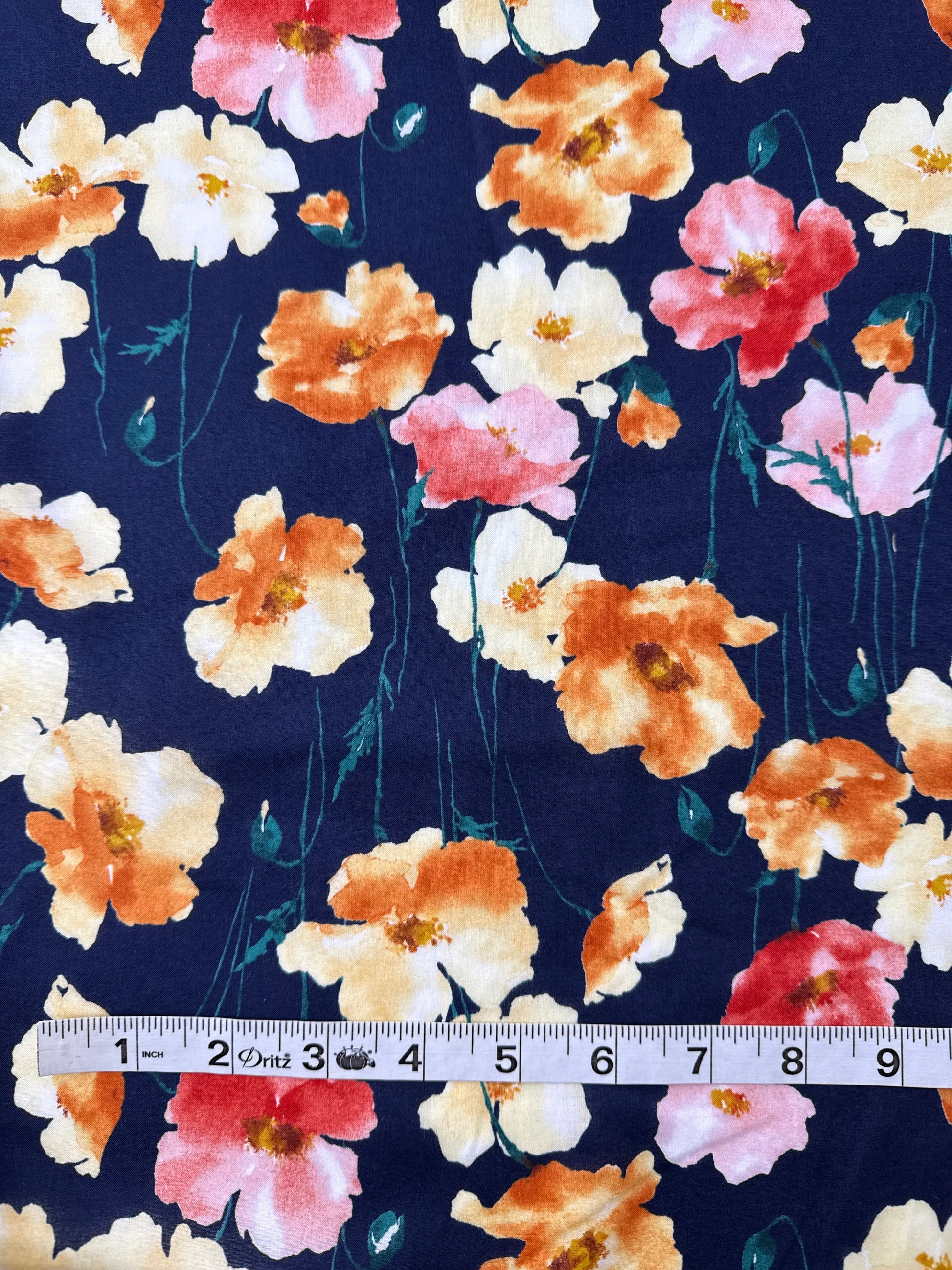 A close-up of floral fabric with a navy blue background and large watercolor-style flowers in peach, cream, pink, and coral. A white measuring tape is placed vertically along the left side for scale.