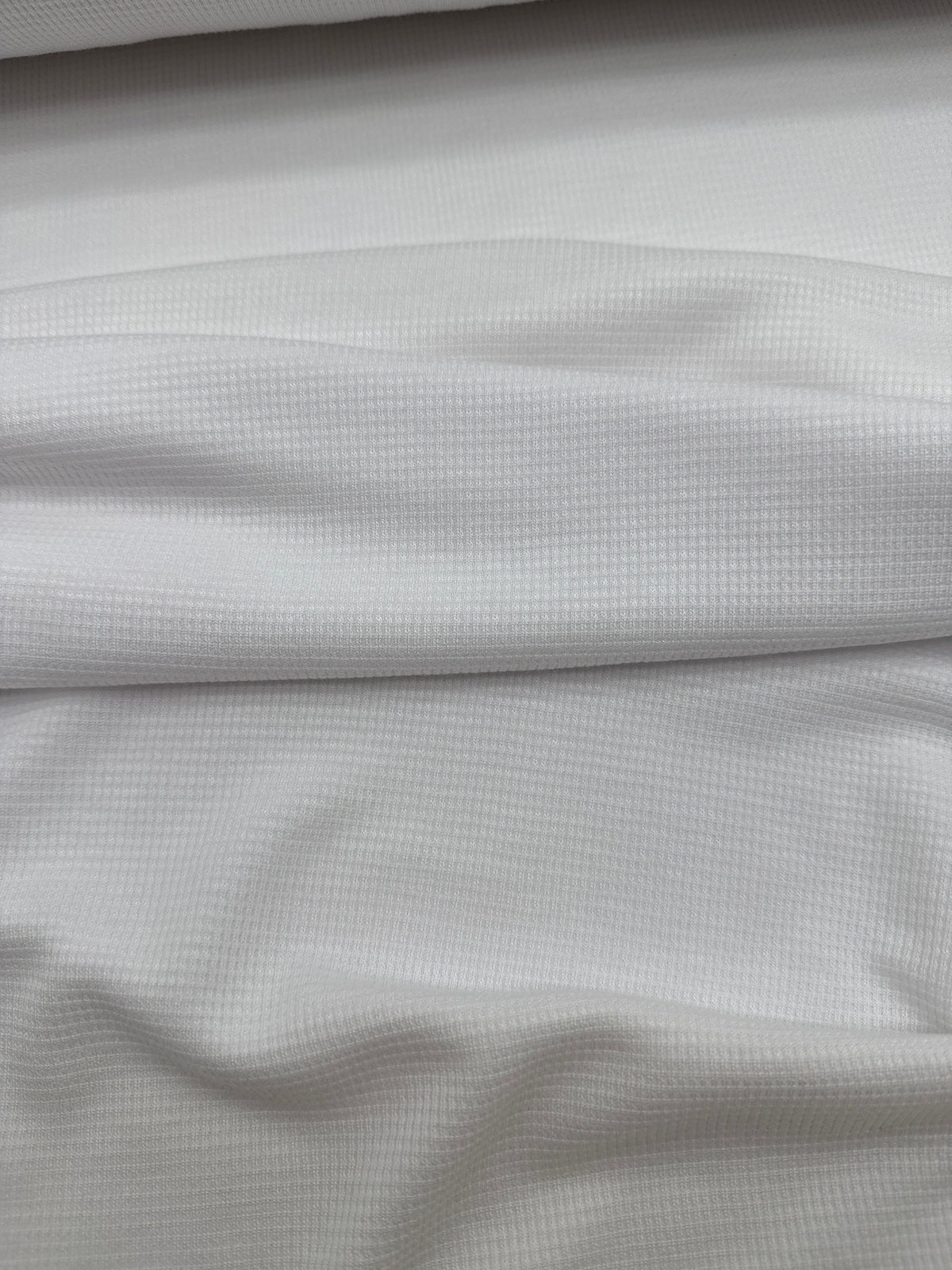 A close-up of white textured fabric with a fine waffle or grid-like weave, shown with soft folds and shadows.