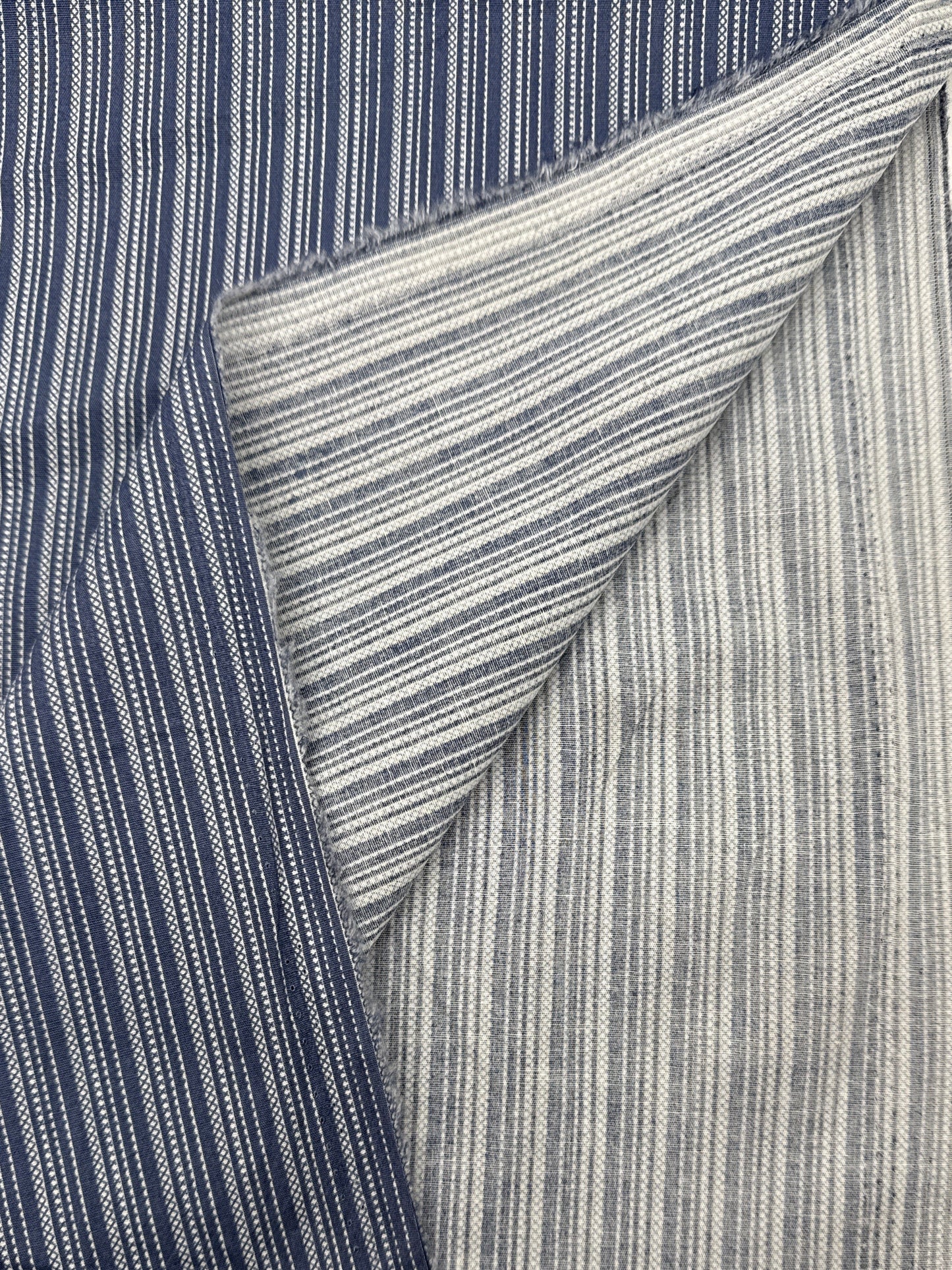 A close-up of striped fabric shown in two tones: a dark navy blue side with thin white vertical stripes, and a lighter gray side with similar white stripe detailing. The fabric is folded to show both sides and has a frayed edge.