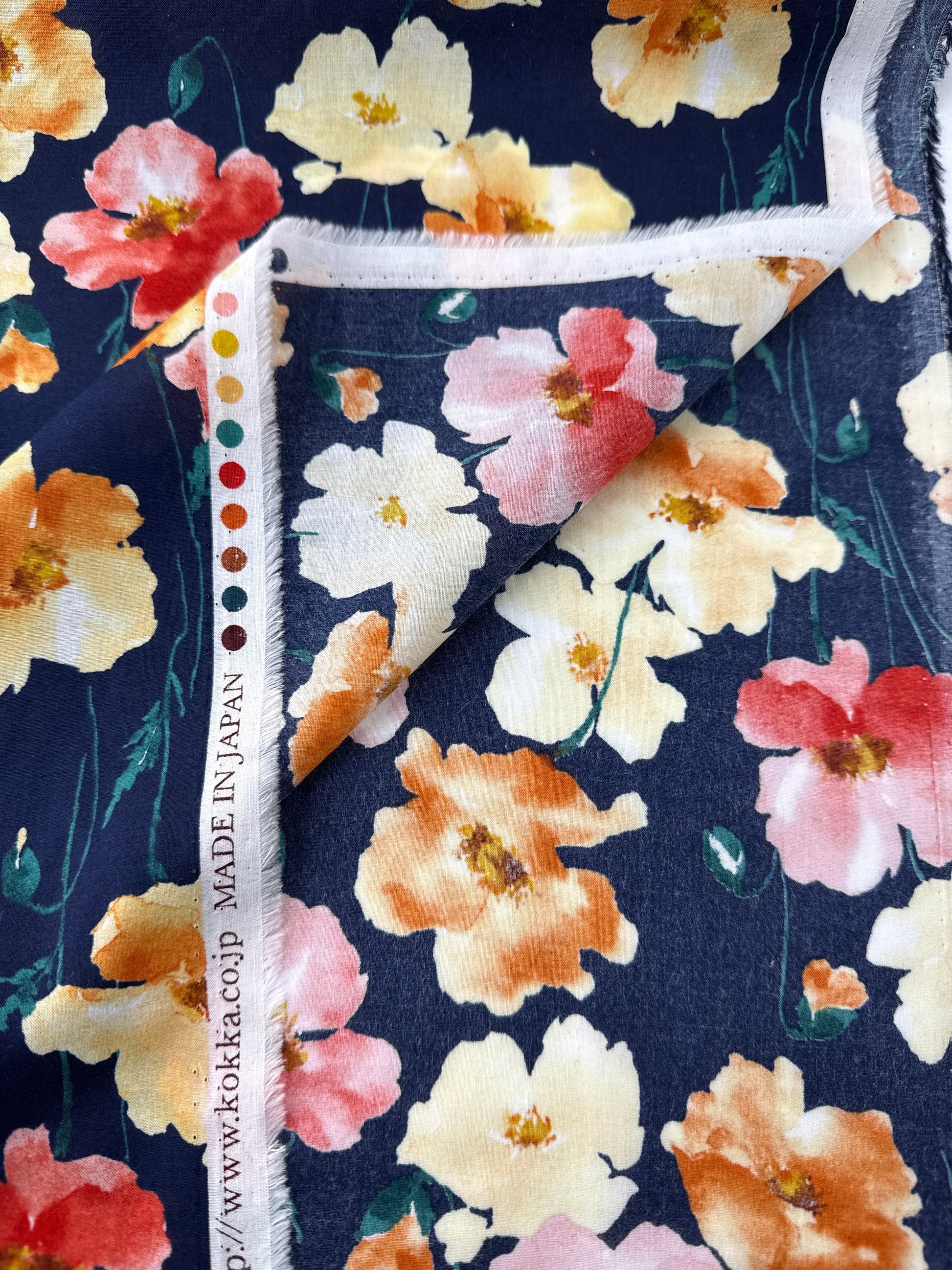 A close-up of a floral fabric with a navy blue background and large watercolor-style flowers in cream, peach, pink, and orange, folded over to show the white frayed edge and printed brand text.