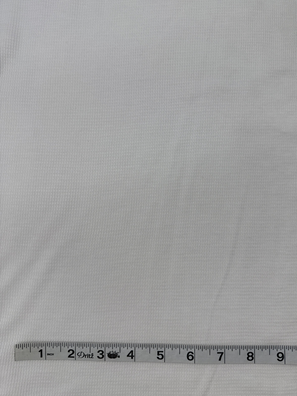 White textured fabric shown close up with a measuring tape ruler along the bottom edge for scale.