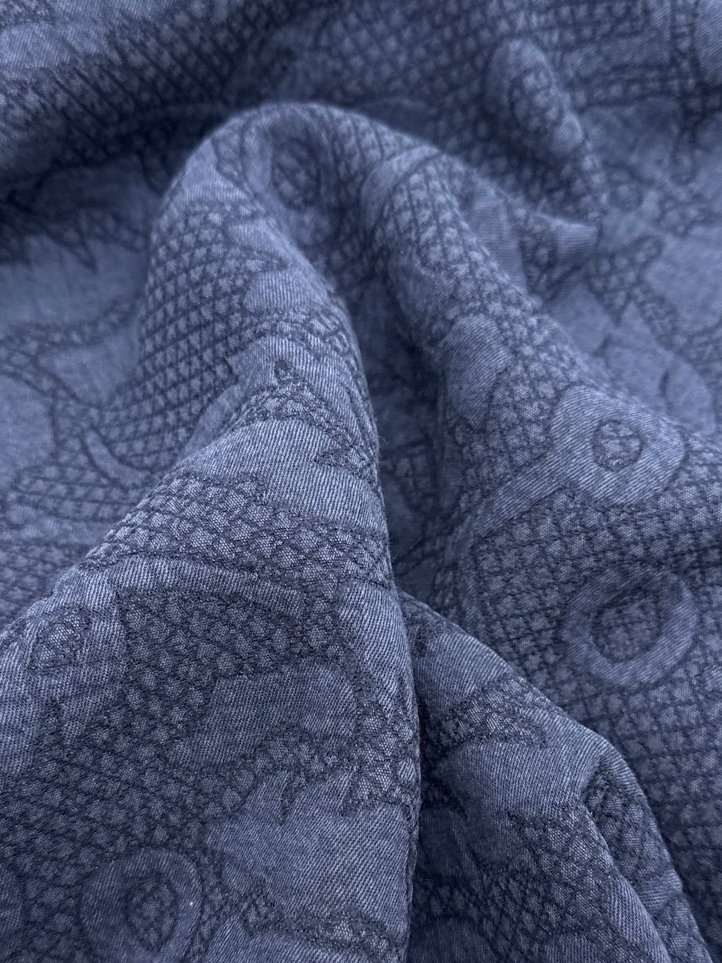 A close-up of draped blue fabric with a tonal repeating floral and geometric pattern, partially covered by white paper edges.
